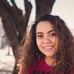 Portrait of a smiling young woman with curly hair in a sunny outdoor setting.